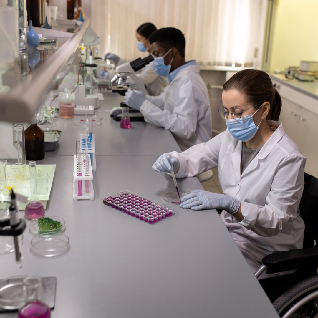 Scientists working in a laboratory, wearing lab coats, masks, and gloves, using microscopes and handling test tubes with pink liquids.