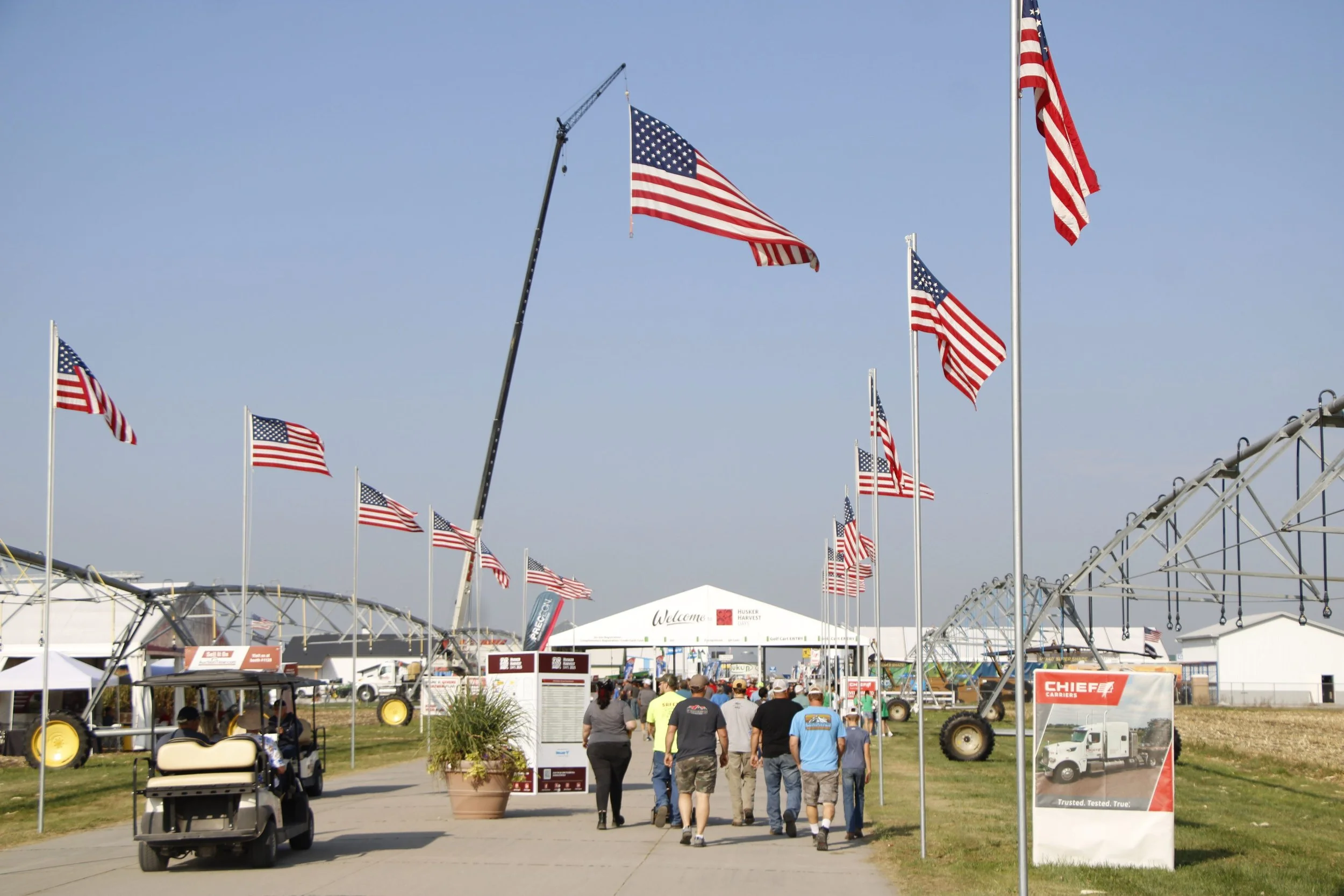 American flags flying over outdoor event tents and a crowd of people walking underneath, with a clear blue sky.