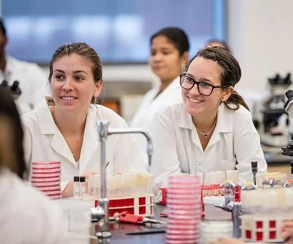 Students in a science classroom wearing lab coats, seated around a lab table with test tubes, and smiling.