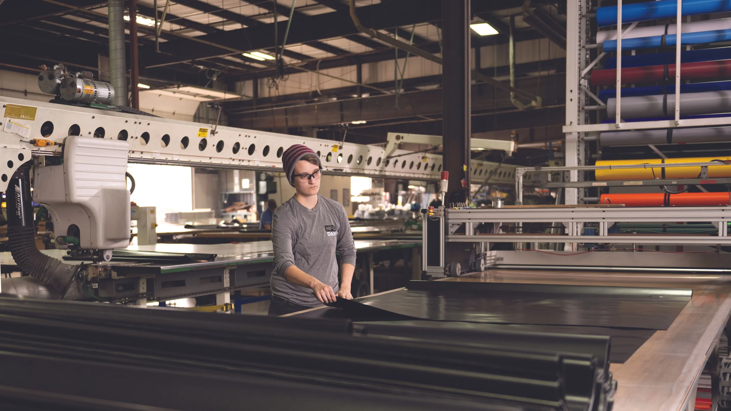 A person wearing glasses and a gray long-sleeve shirt operating machinery in a manufacturing or industrial workshop with rolls of material and equipment around.