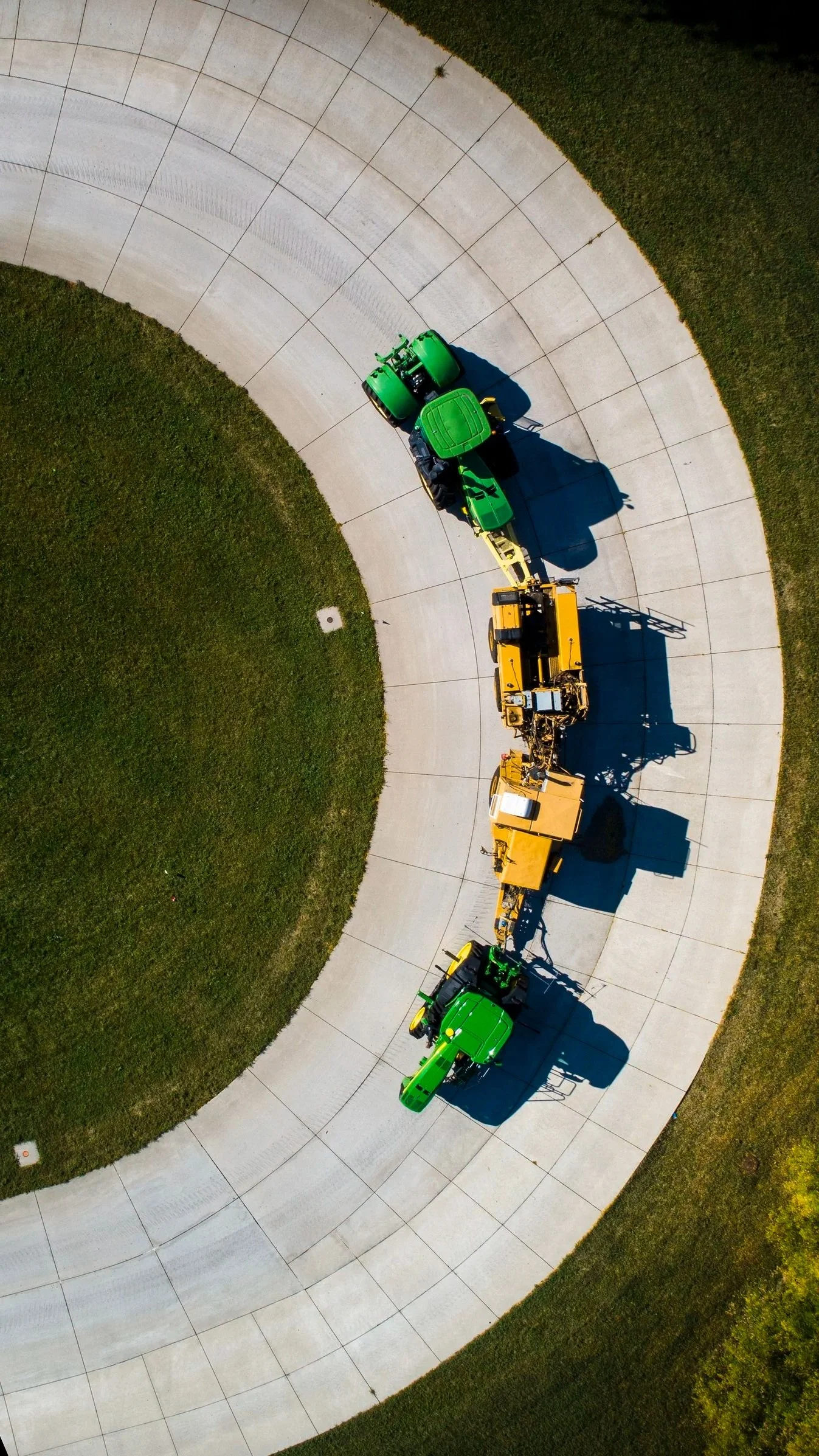 Aerial view of green and yellow construction and farming machinery parked in a curved formation on a concrete surface, with green grass on either side.