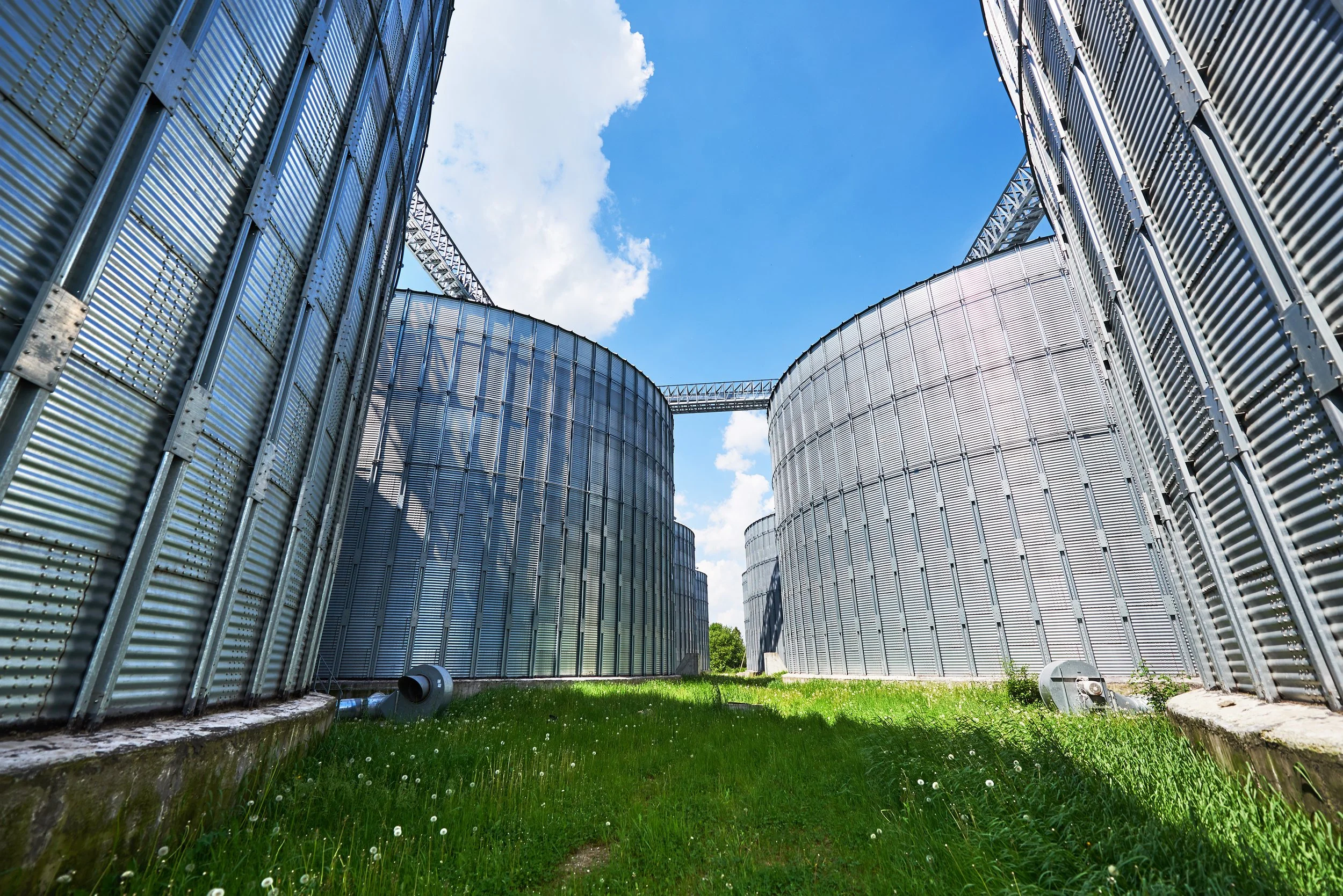 A low-angle view of large industrial grain silos made of metallic material, with a blue sky and white clouds above.