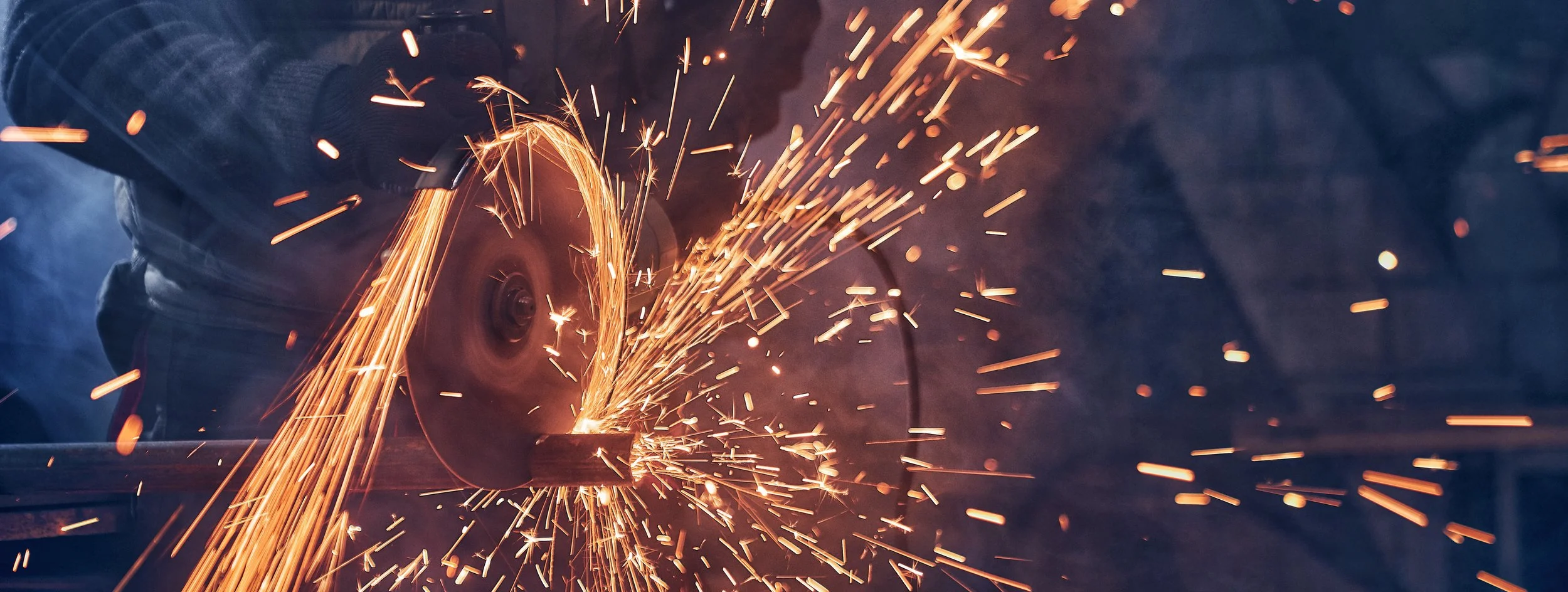 A worker using an angle grinder cuts through a piece of metal, producing a shower of bright sparks.