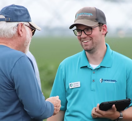 Two men shaking hands outdoors, one holding a tablet, both wearing hats and glasses, with a grassy field and cloudy sky in the background.