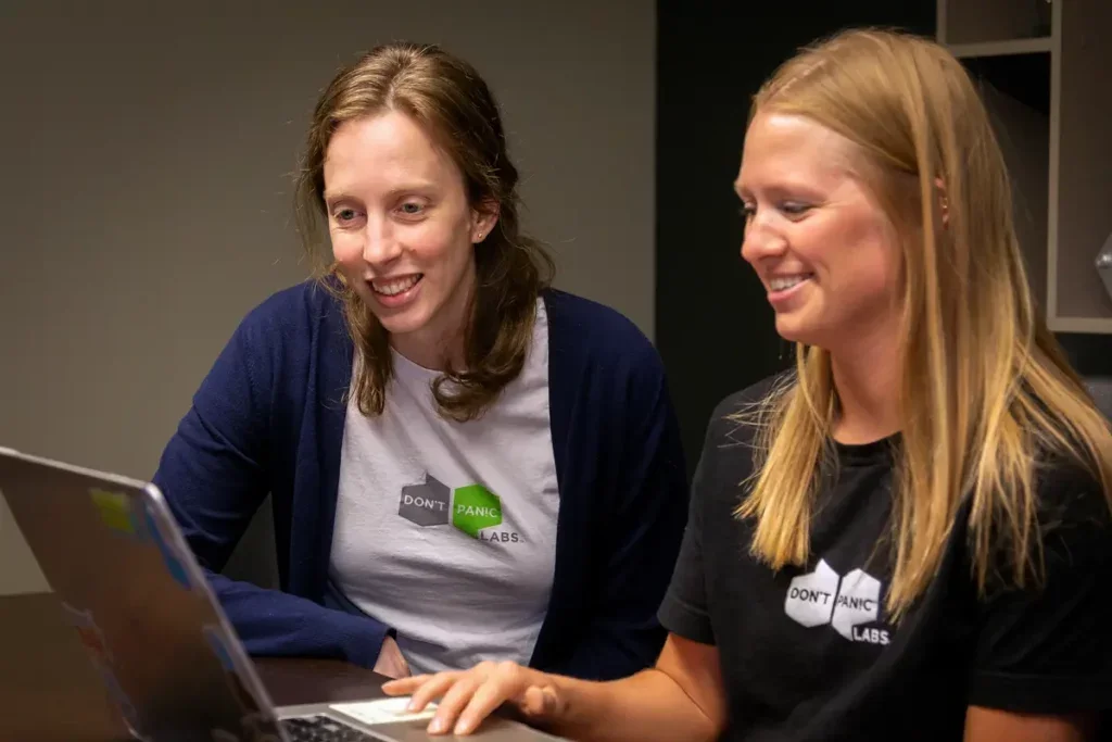 Two women sitting at a table, looking at a laptop, smiling, wearing T-shirts with the print 'DON'T PANIC LABS'.