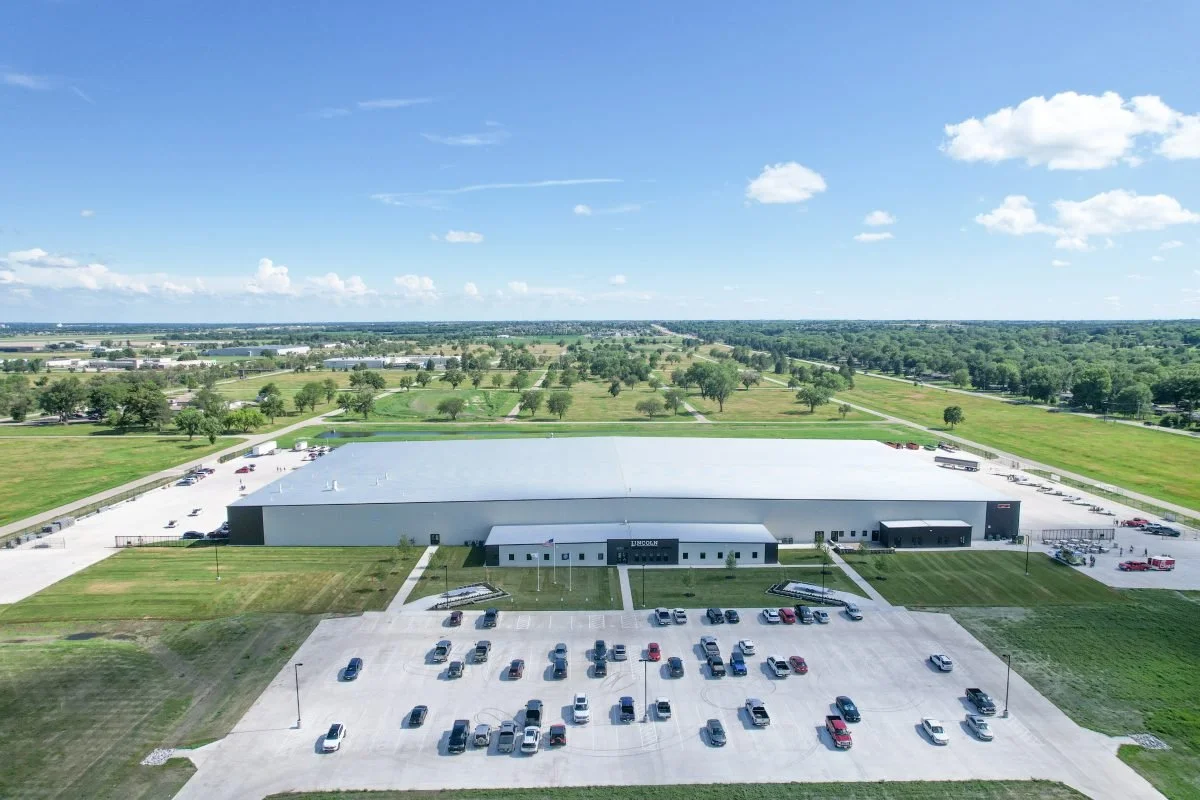 Aerial view of a large industrial warehouse with a parking lot in the foreground, surrounded by green fields and trees under a partly cloudy sky.