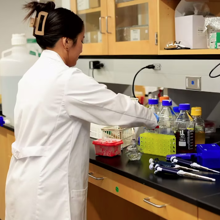 A scientist working in a laboratory, handling glass bottles and equipment on a black countertop with wooden cabinets and electrical outlets in the background.
