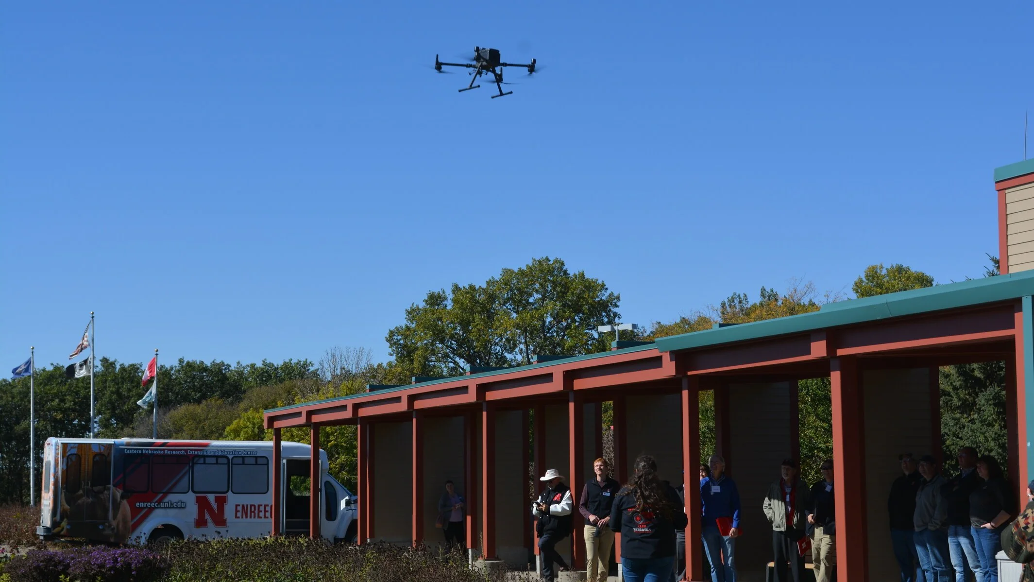 A drone flying in the sky above a group of people standing under a shelter, with flags and a bus nearby, and trees in the background on a clear day.