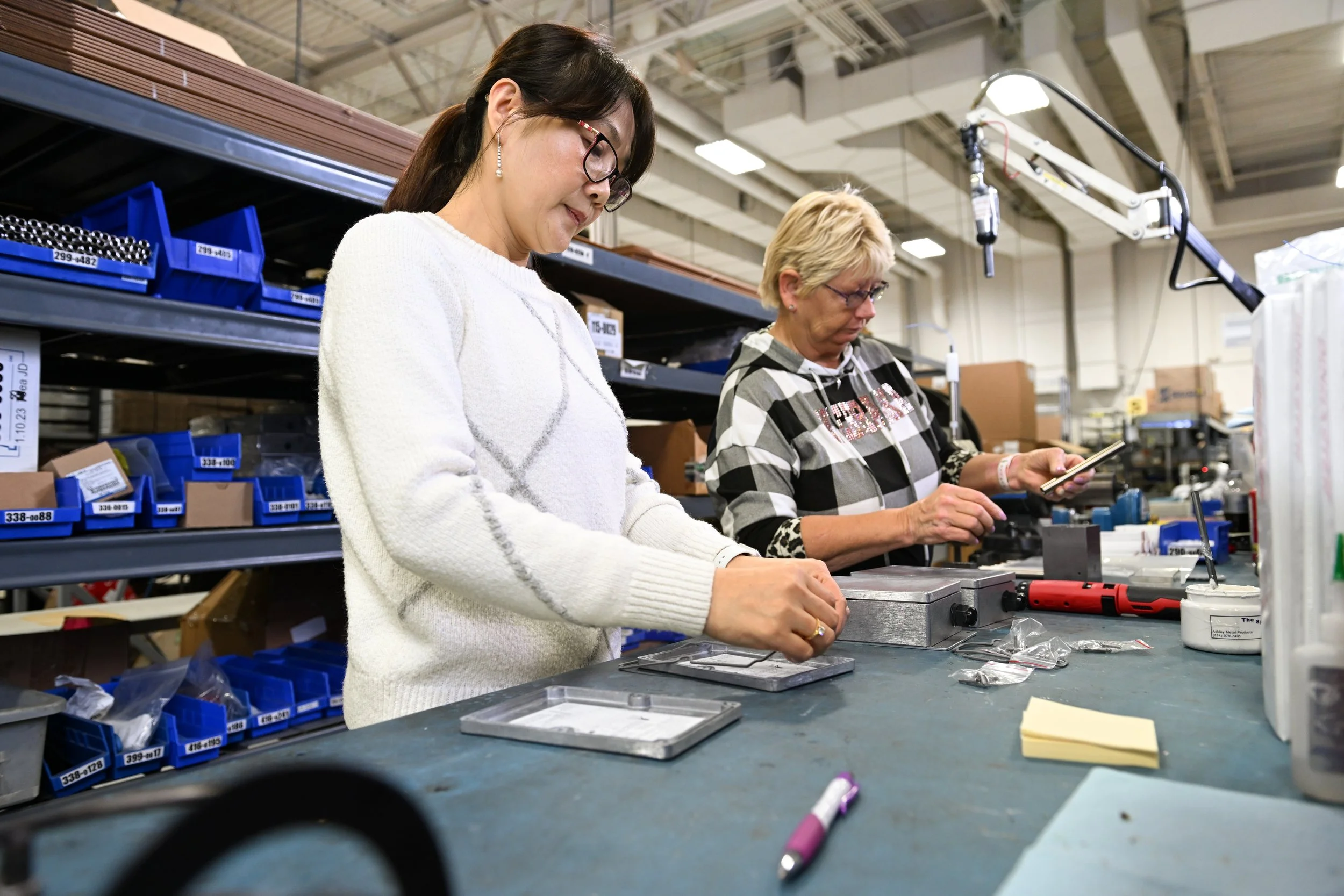 Two women working at a workstation in an industrial or manufacturing setting, surrounded by blue bins filled with small parts and tools.