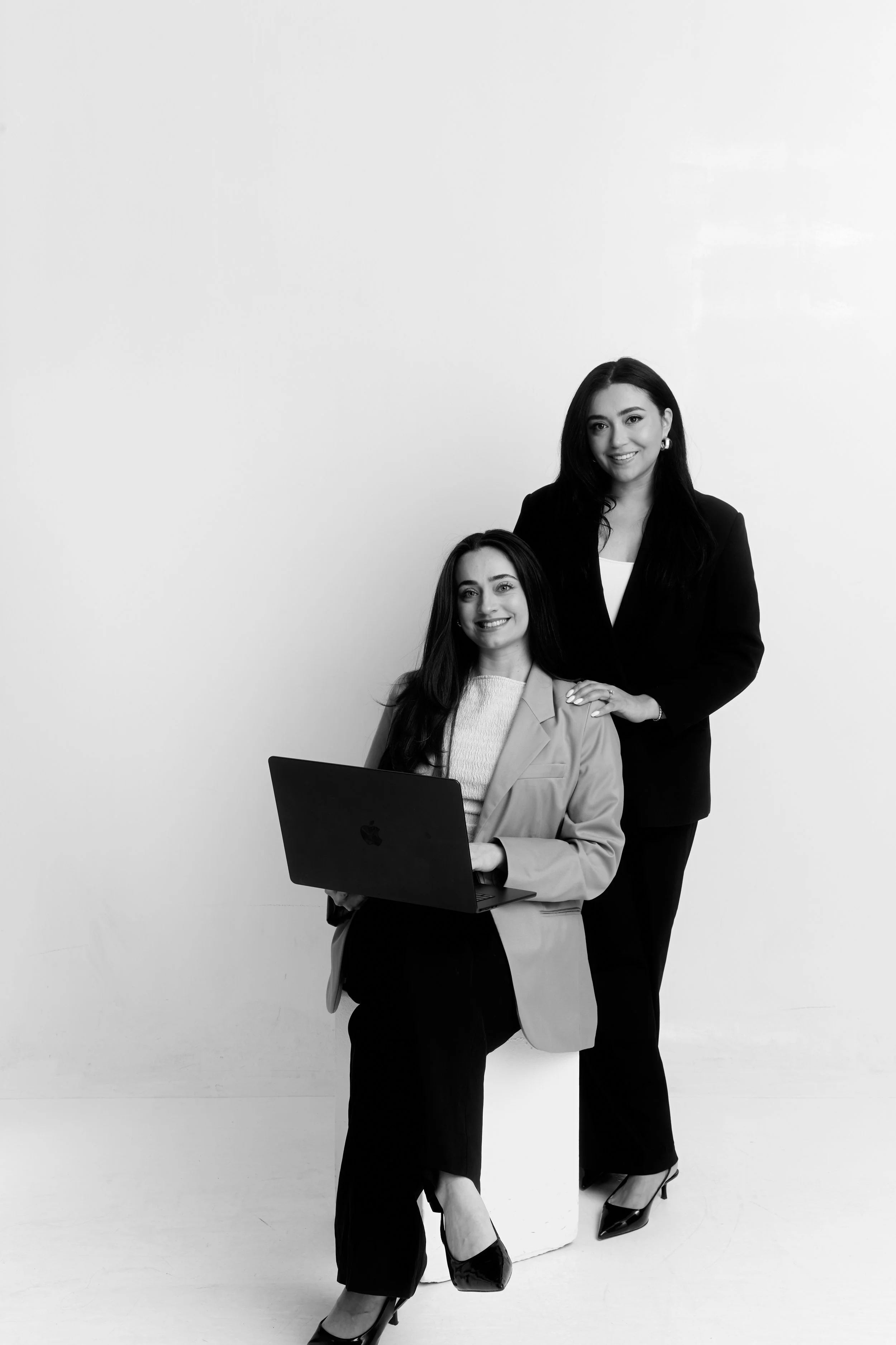 Two women dressed in business attire, one sitting with a laptop and the other standing behind her, both smiling, against a plain white background.