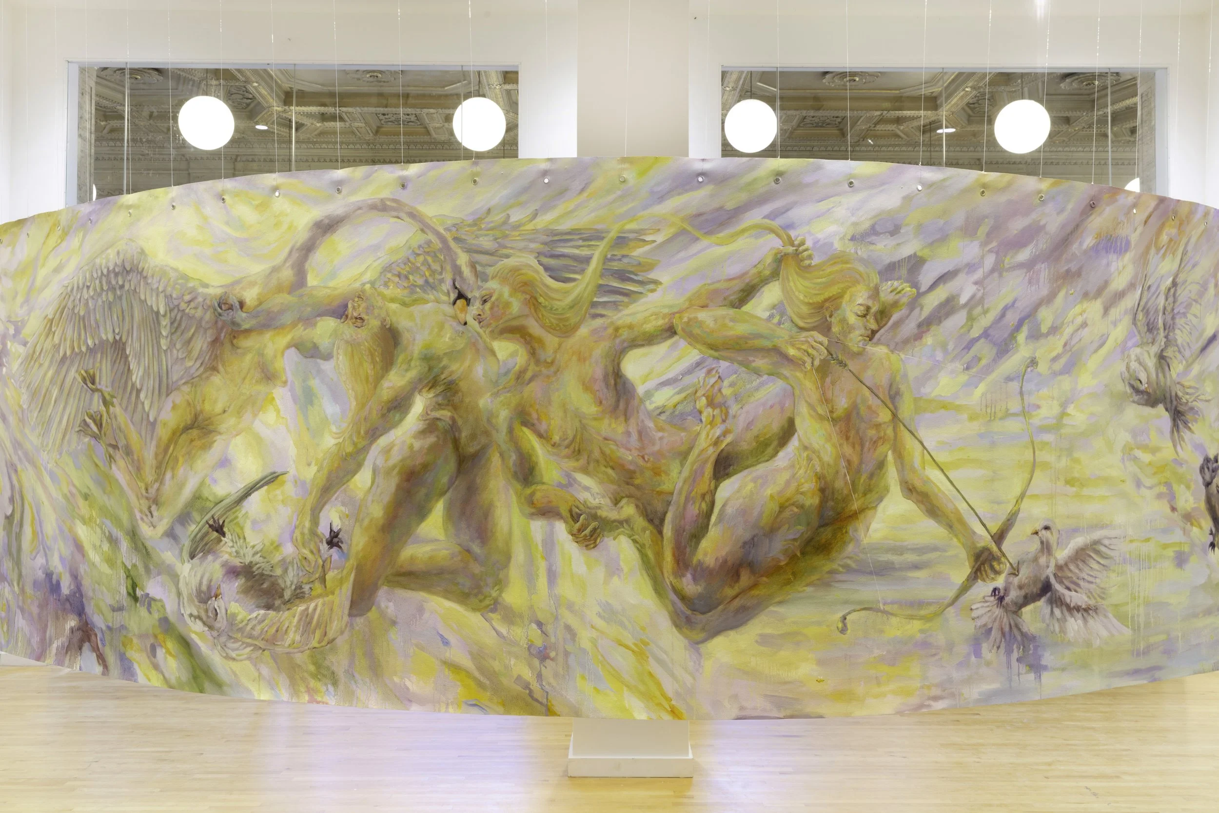A large painting depicting mythological figures and flying creatures in soft pastel colors, set in an indoor gallery with ornate architectural details and round ceiling lights.