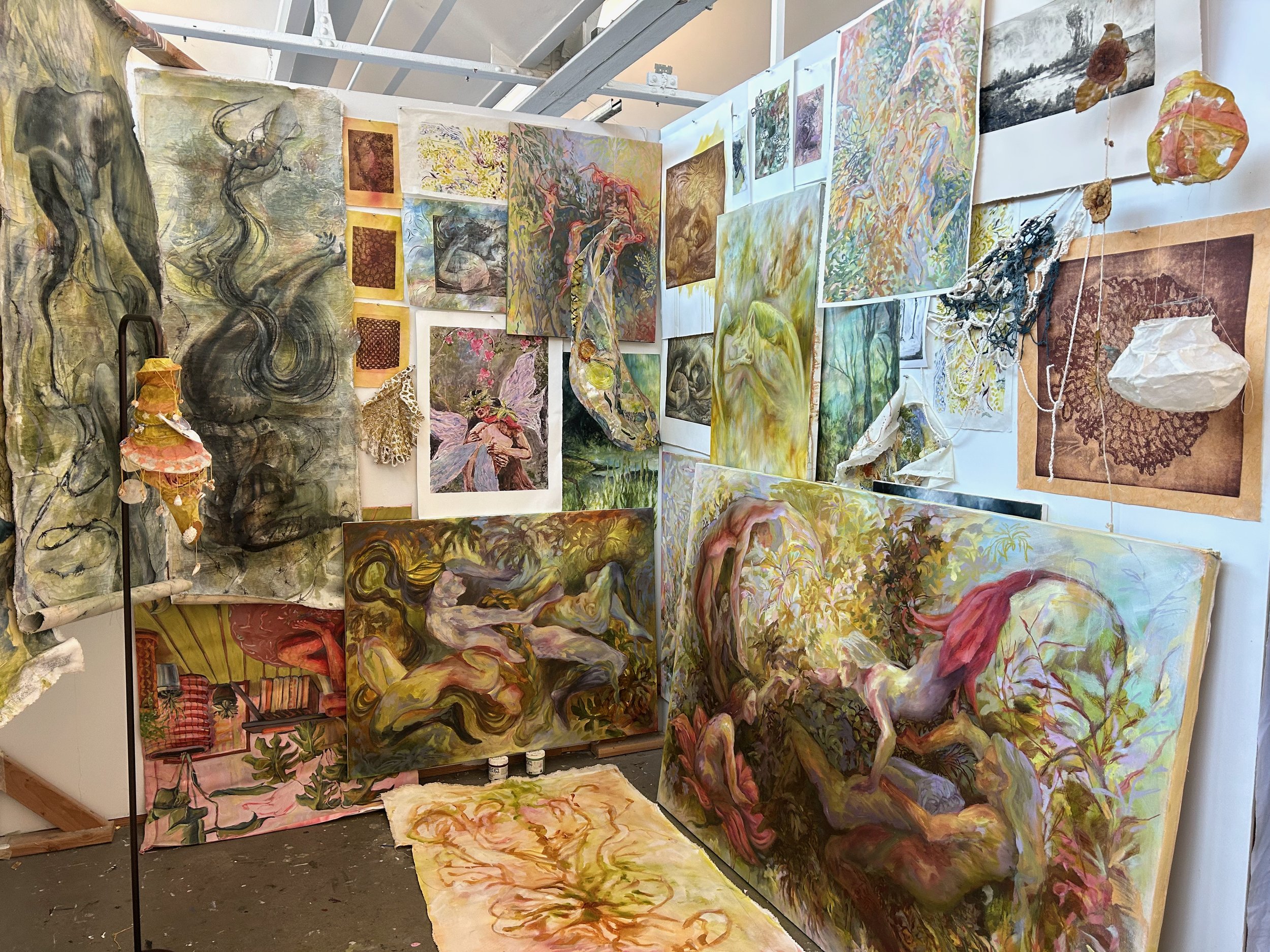 An art studio with various paintings and sculptures, including abstract, figurative, and fantasy-themed artworks, displayed on white walls and standing on the floor, with some hanging and others leaning.