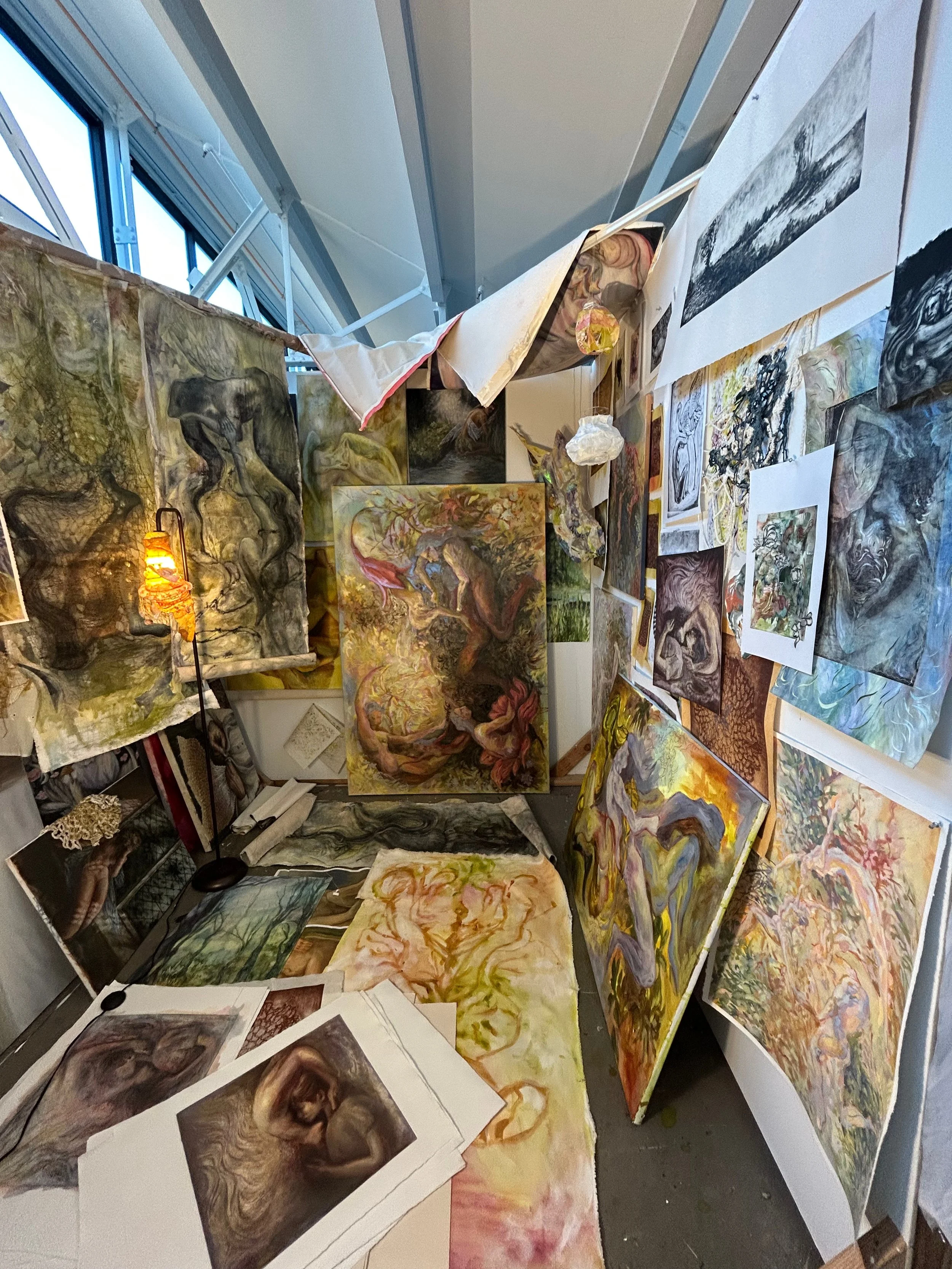 An art studio wall filled with various colorful abstract and figurative paintings, sketches, and art supplies, with some artwork leaning against the wall and others hanging or taped on the surfaces.