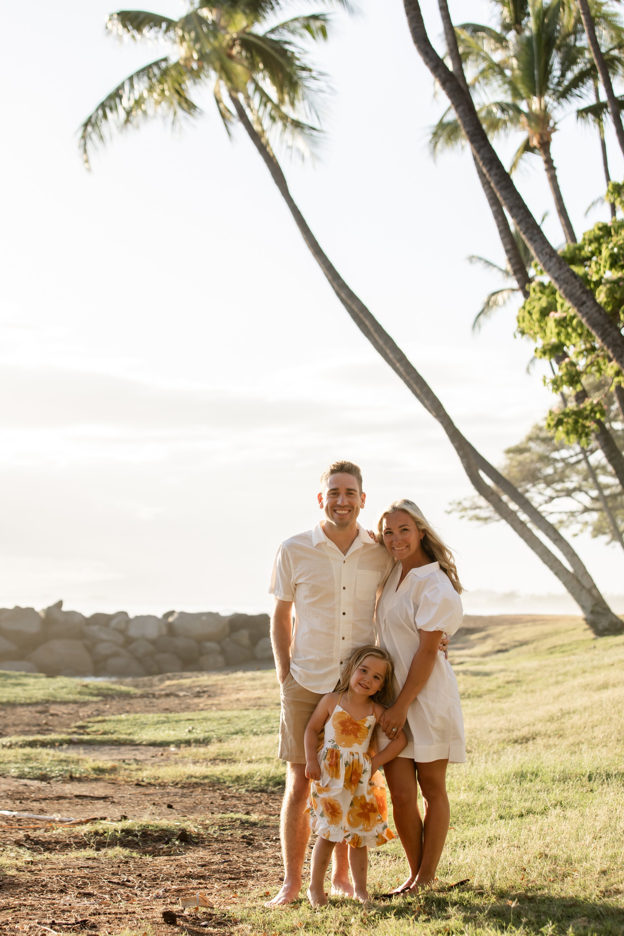 A happy family of three standing outdoors on a grassy area near the beach, with palm trees and a rocky shoreline in the background during sunset.