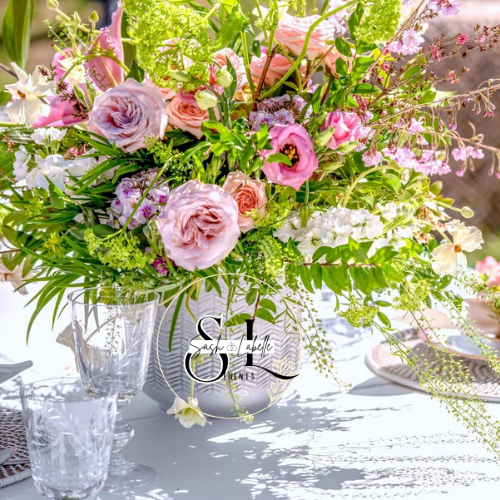 A large flower arrangement in a vase with pink, white, and purple flowers and green foliage, placed on a white table with glassware and table settings.