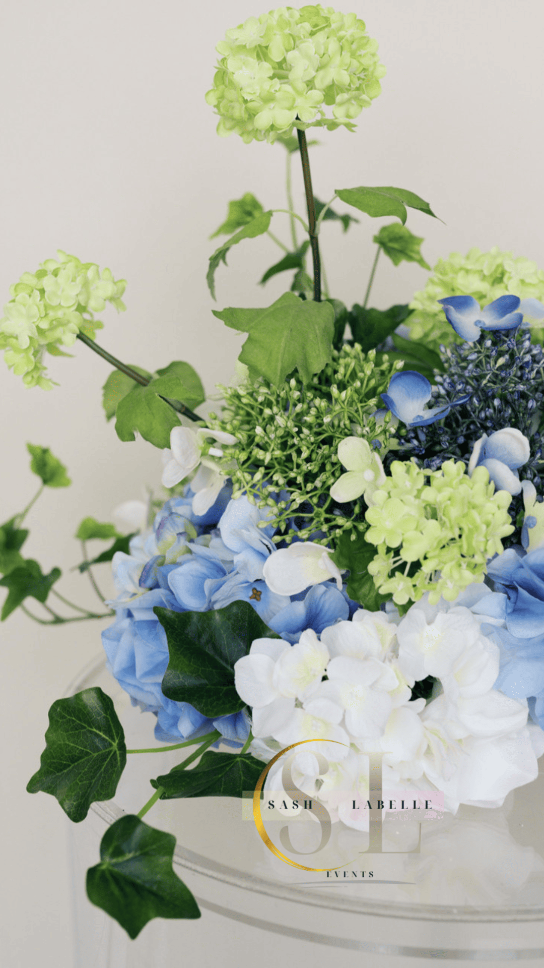 A floral arrangement with white, blue, and light green hydrangeas, and green leaves in a transparent vase with the logo and text 'Sash Labelle Events'.