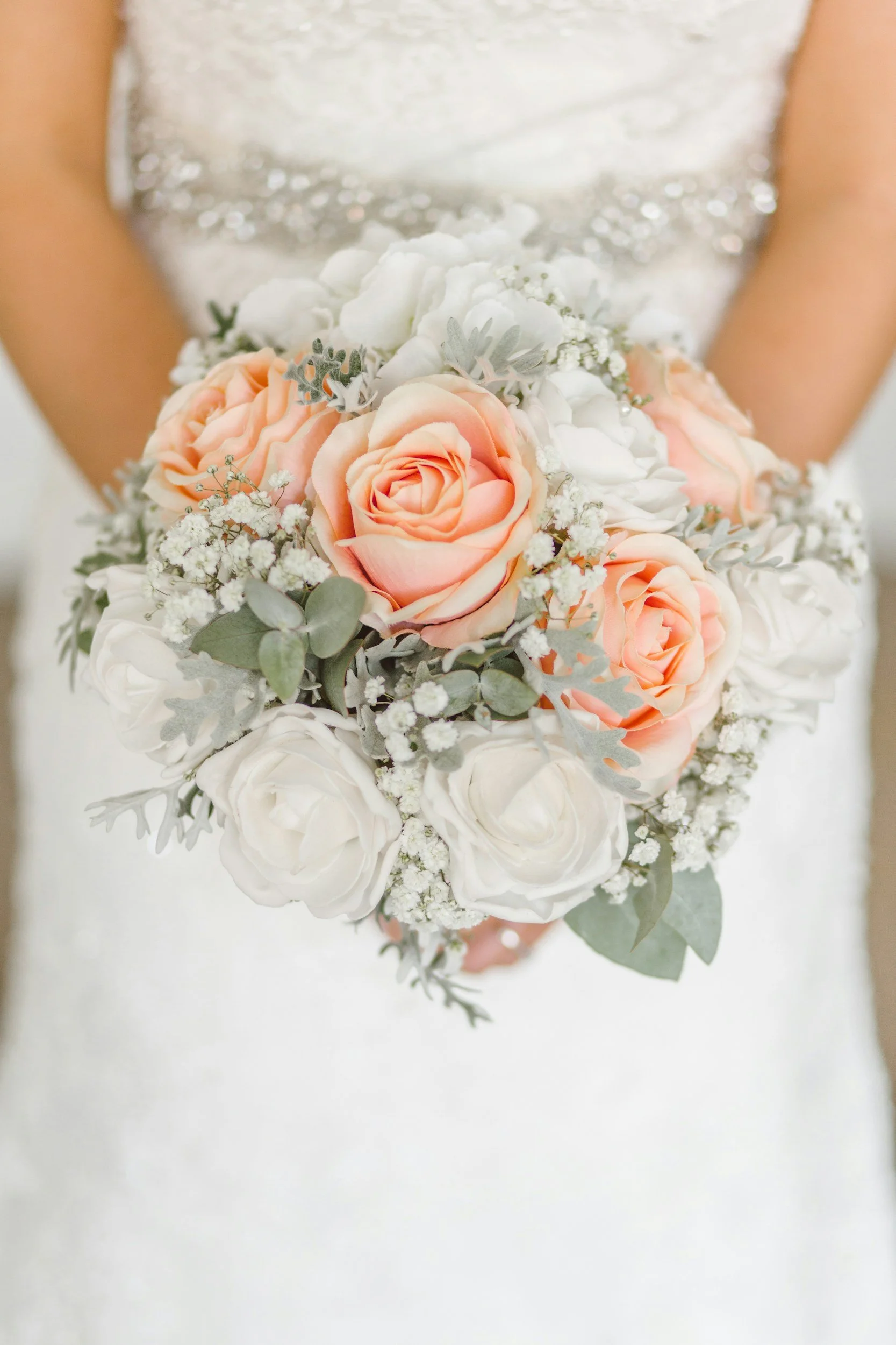 Close-up of a bridal bouquet with peach roses, white flowers, and green leaves, held by a bride in a sparkling, silver dress.