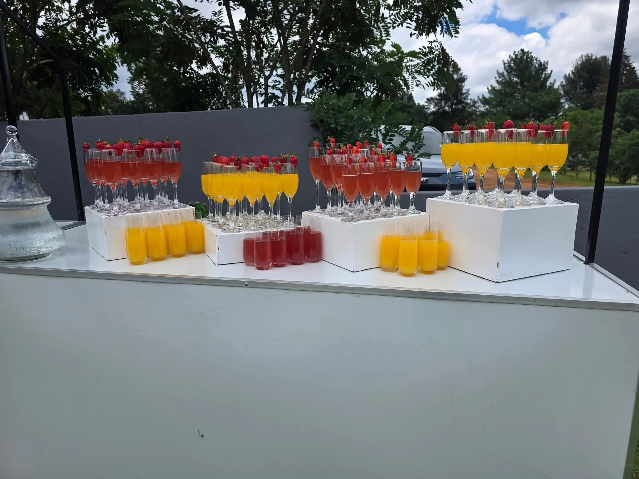 Multiple glasses filled with orange and pink beverages, garnished with strawberries, placed on white boxes. A glass pitcher of water is on the left side. The background has trees and a blue sky with clouds.