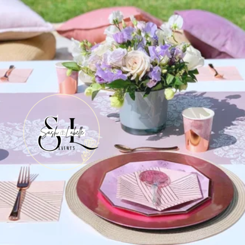 Table set for a special event with a large flower centerpiece, pink and purple accents, and elegant tableware, including a plate, napkin, utensils, and cups.
