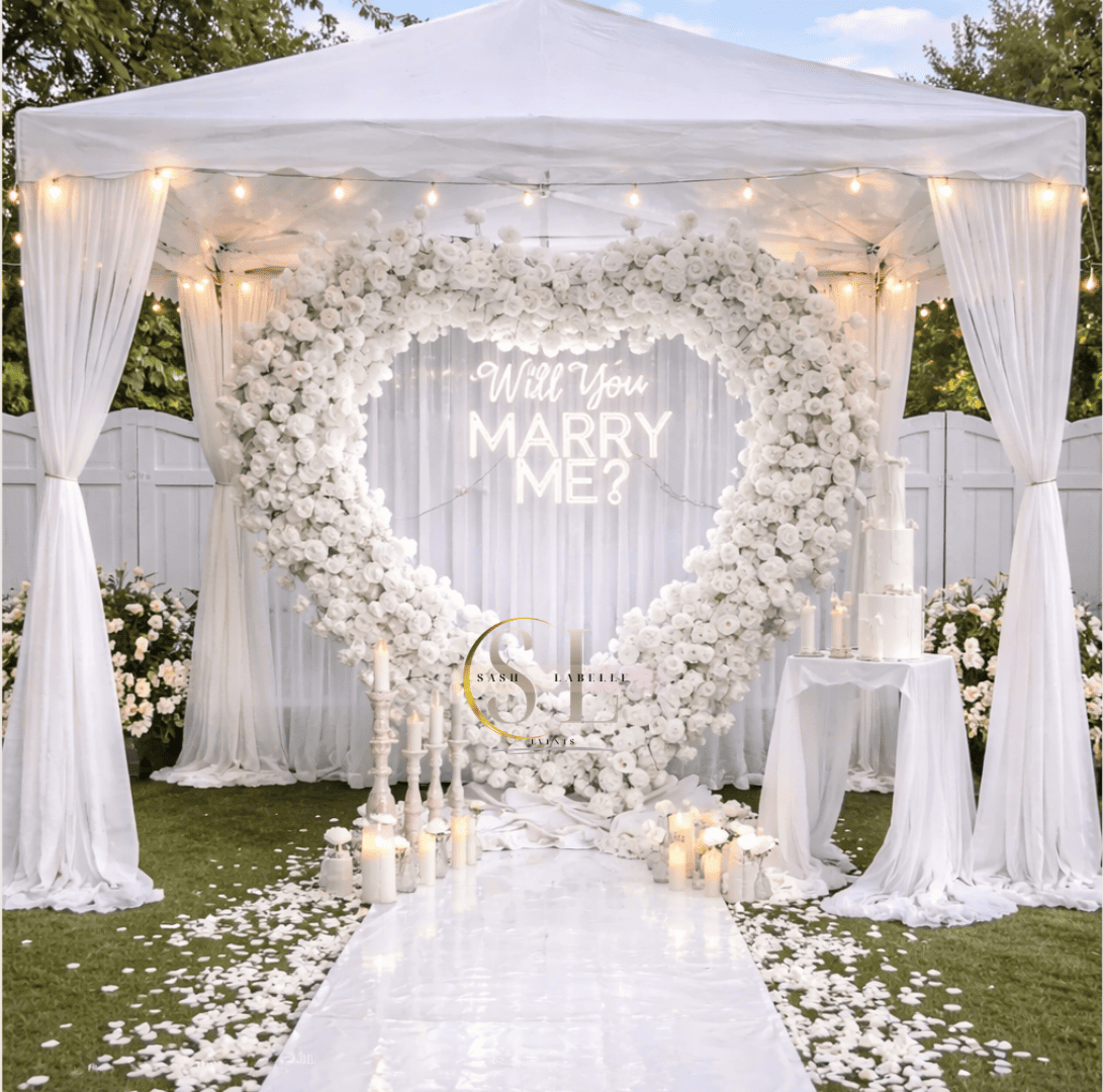 White wedding arch decorated with white flowers, draped white curtains, and string lights. A neon sign asking "Will You MARRY ME?" is centered within the arch. Candles and a wedding cake are on white tables nearby, with flower arrangements on the ground. The setting is outdoors with a white fence in the background.