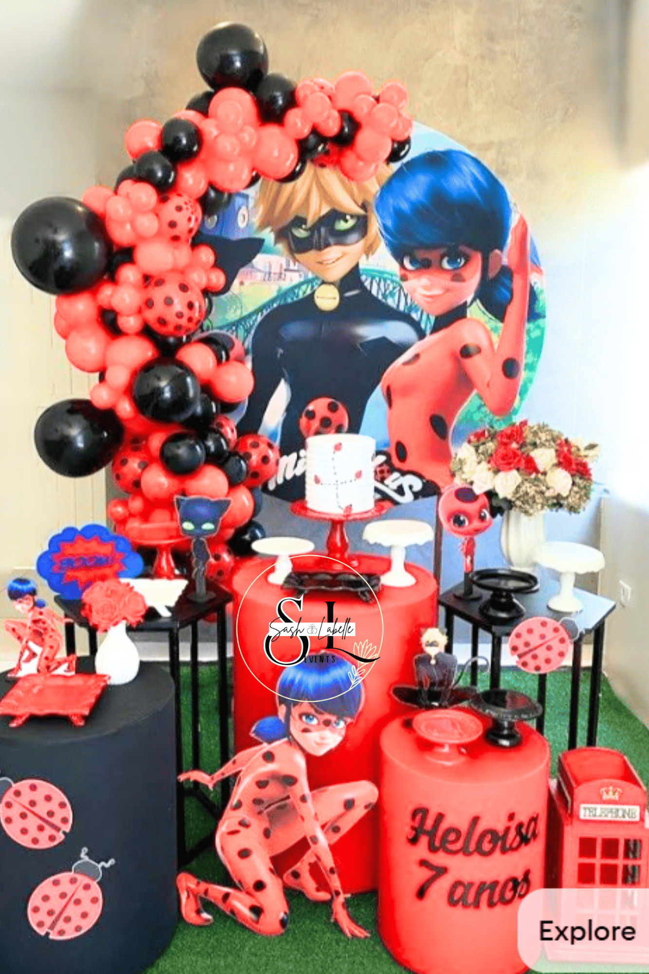 A themed birthday party setup featuring decorations from Miraculous Ladybug, including balloons, cutouts, and table decor in red, black, blue, and white. There is a large backdrop with characters Ladybug and Cat Noir, a cake, and various themed decor