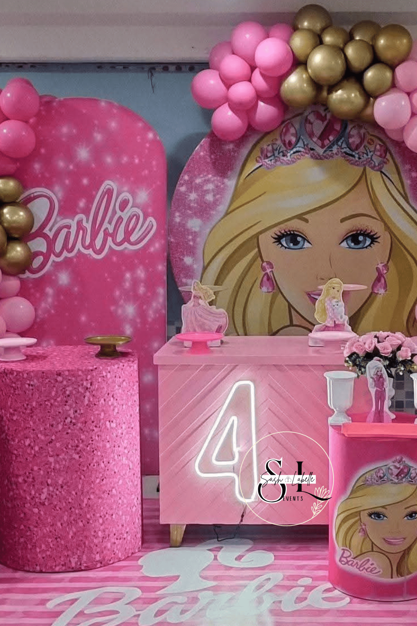 Barbie-themed birthday party decorations featuring pink and gold balloons, a large cutout of Barbie's face with a tiara, a pink glittered table, a pink table with small figurines of Barbie, and a pink backdrop with the word "Barbie" written on it.