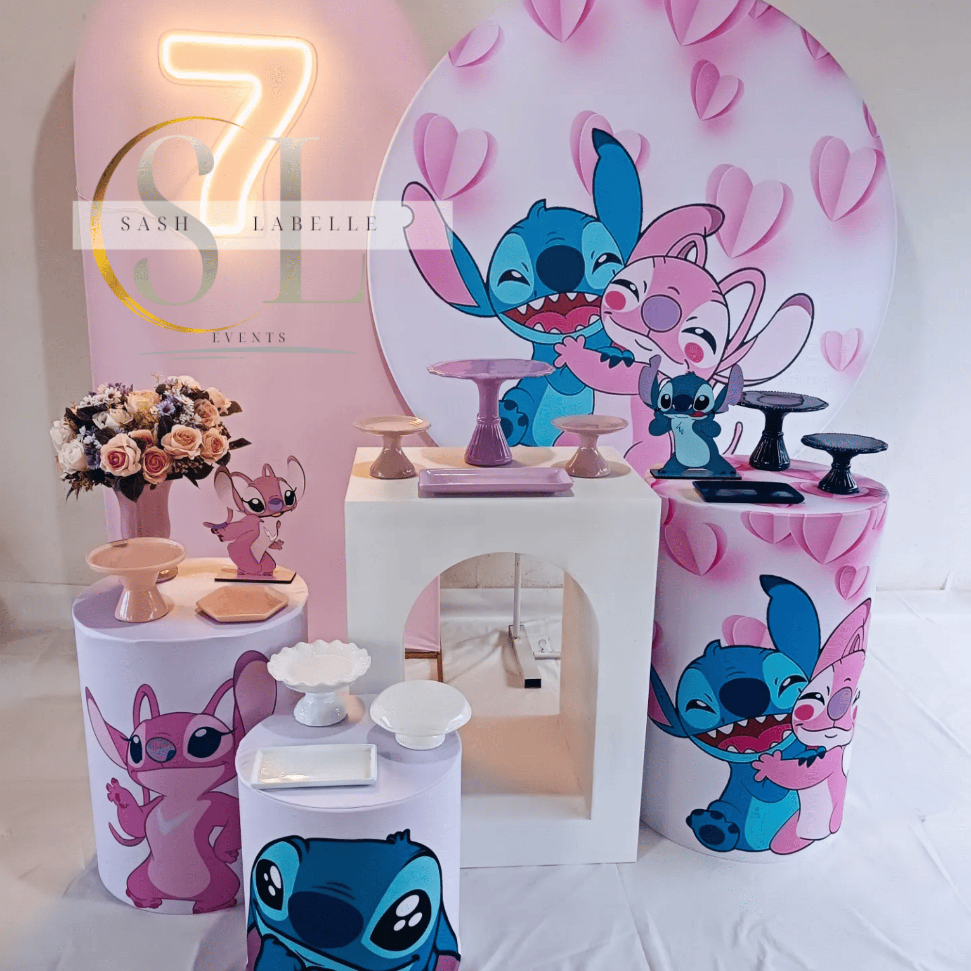 Decorative setup with cartoon characters from the show 'Lilo & Stitch,' pink heart-shaped paper decorations, a bouquet of pink roses, and multi-tiered pastel-colored dessert stands in front of a pink backdrop with the words 'Sash LAbelle Events.'