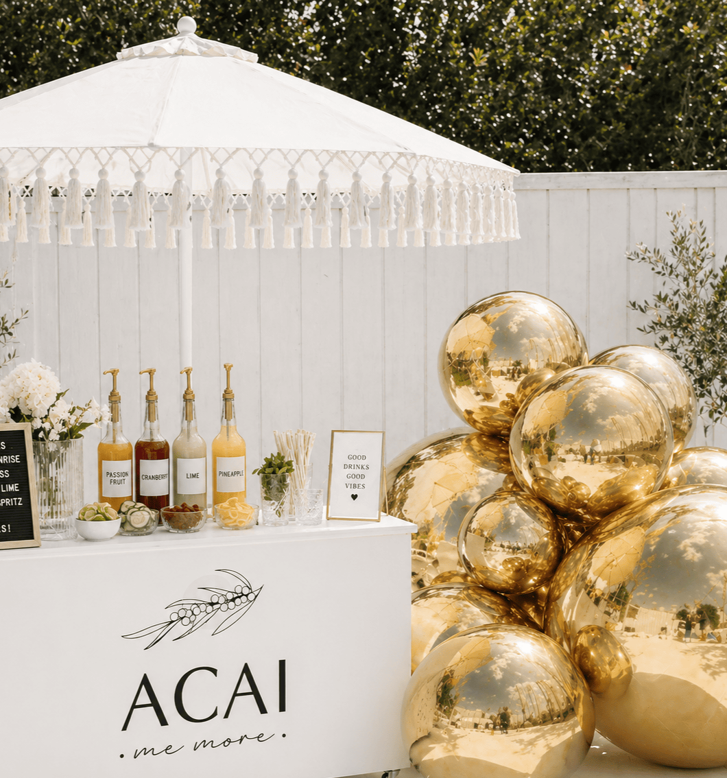 A white outdoor bar setup with a large white parasol, gold reflective balloons, bottles of flavored liquids, and a sign that says 'ACAI me more'. Small bowls of toppings and a framed sign with a positive message are also visible.