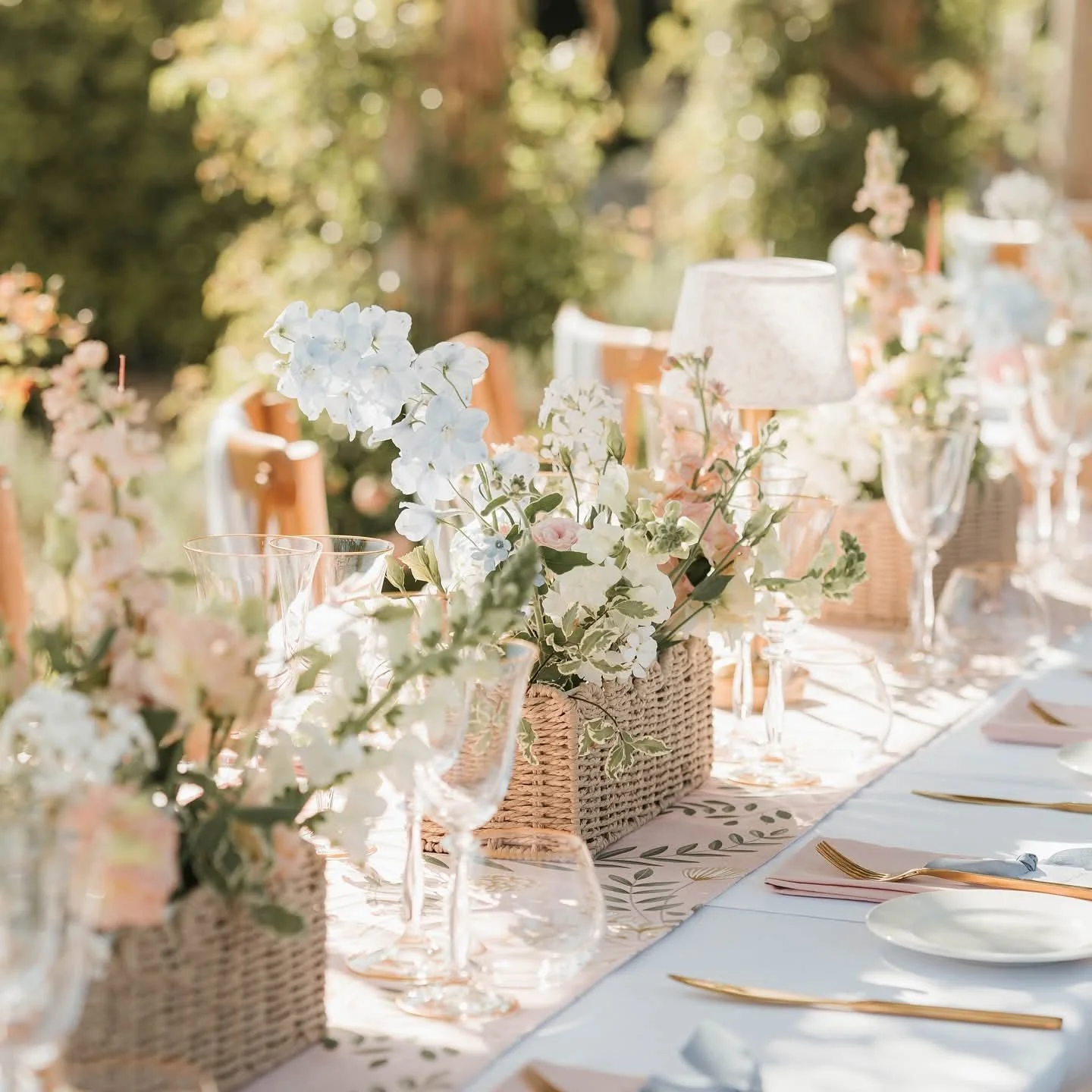 Elegant outdoor table decorated with white and pastel floral arrangements in woven baskets, clear glassware, white plates, and gold utensils, set in a sunny garden.