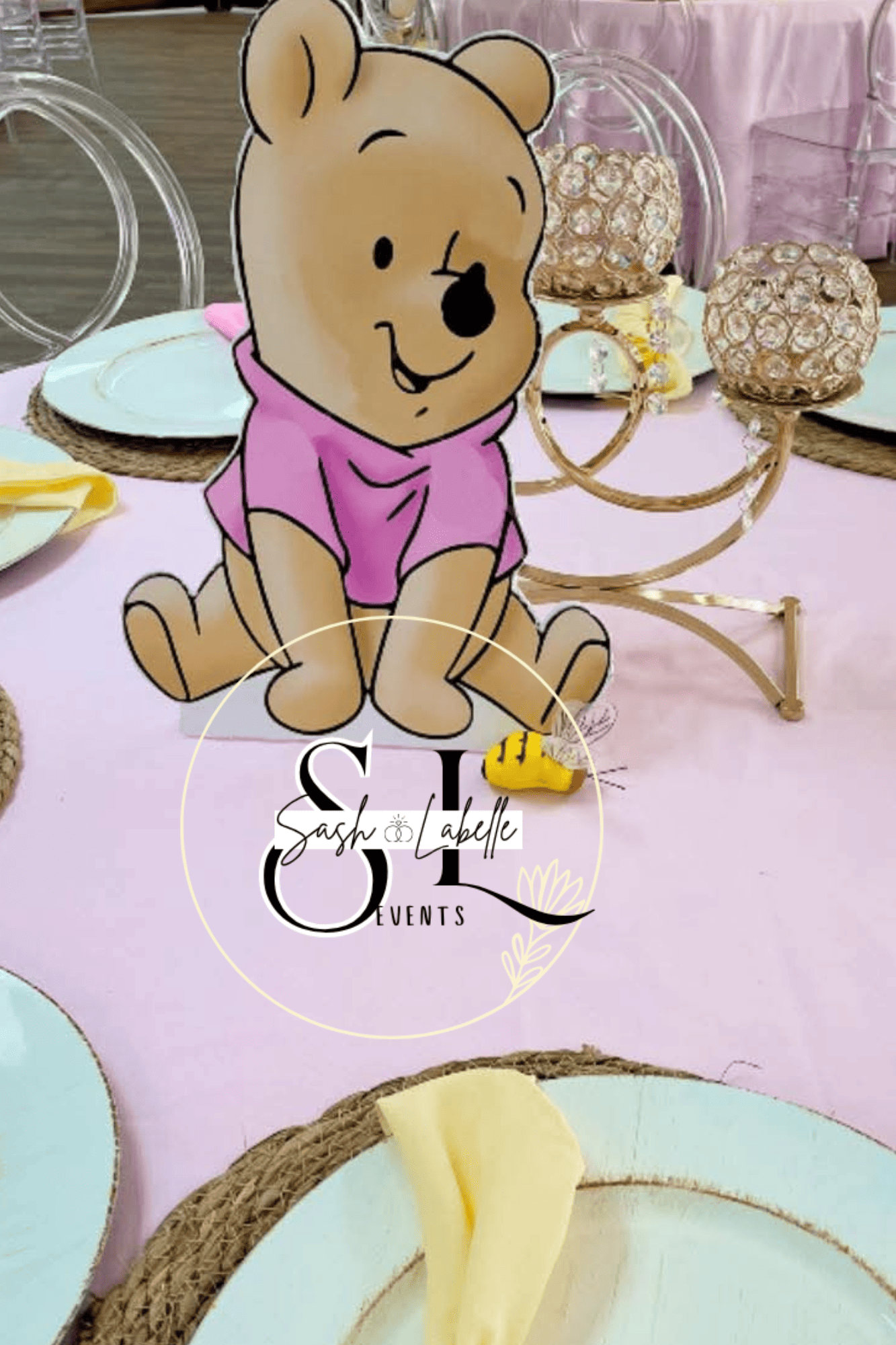 Decorative table setting with a cartoon bear figure, gold candleholders, and pastel-colored plates and napkins, indicating a celebration or event setup.