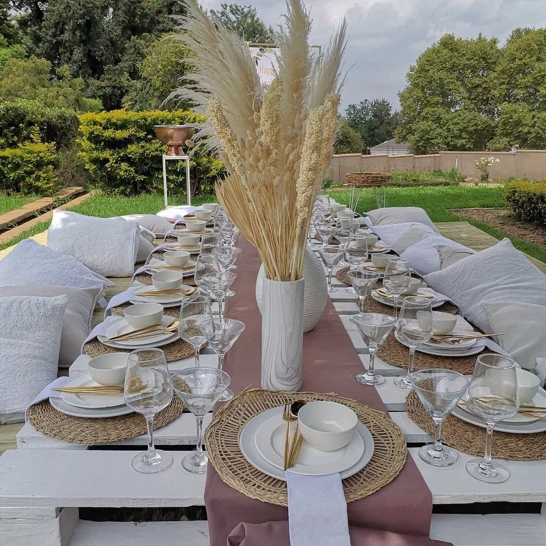 Outdoor dining table set for a gathering with white plates, bowls, gold utensils, clear wine glasses, and woven placemats on a pink table runner, topped with a large white vase filled with dried pampas grass, surrounded by white pillows in a garden s