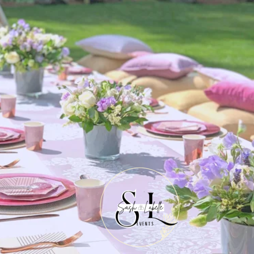 Beautiful outdoor table setting with pink and purple flowers, pink cups, and pastel-colored cushions on the grass.