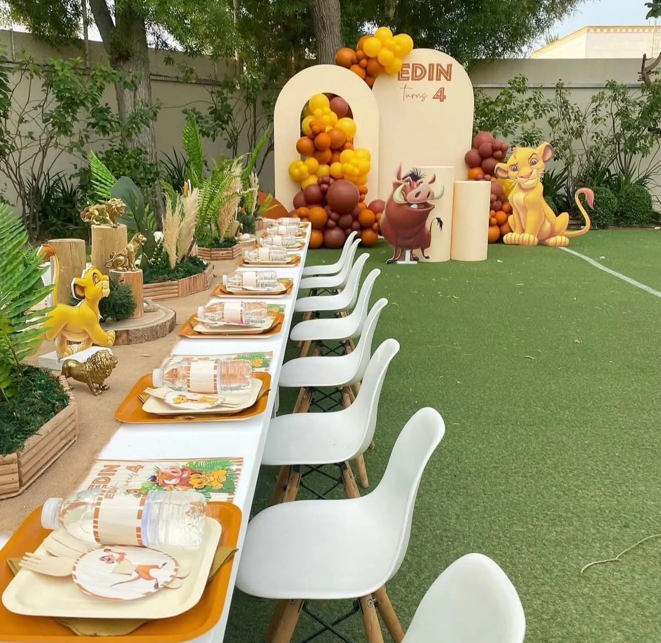 Table set for a birthday party with animal themed decorations, including a backdrop with balloons, cartoon animal characters from The Lion King, and jungle-themed plants, chairs arranged along the table, and a grassy outdoor setting.