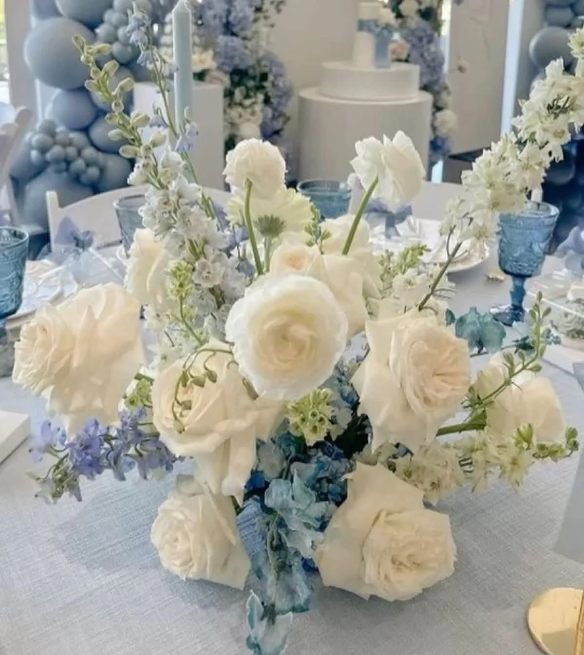 Elegant table centerpiece with white and blue flowers including roses, hydrangeas, and delphiniums, set on a white tablecloth with blue glassware and candles, in a beautifully decorated event setting.