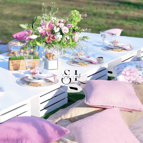 Outdoor table set with floral centerpiece, pink and purple pillows, white plates, glasses, and cutlery on a white tablecloth, with a grassy field in the background.