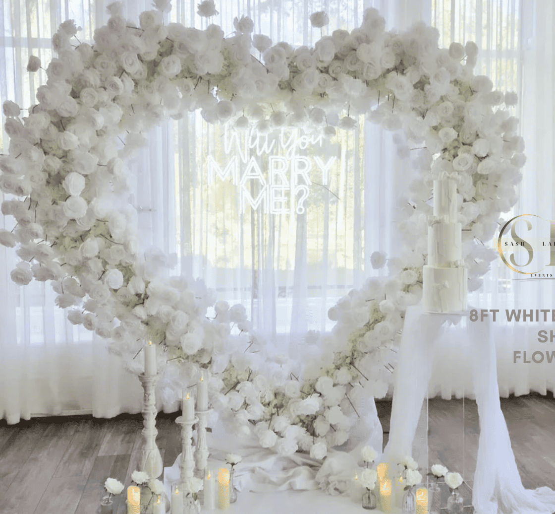 A white heart-shaped floral arch made of roses and flowers, decorated with candles in glass jars and a neon sign that reads 'Will You Marry Me?' in the background, suggesting a marriage proposal setup.