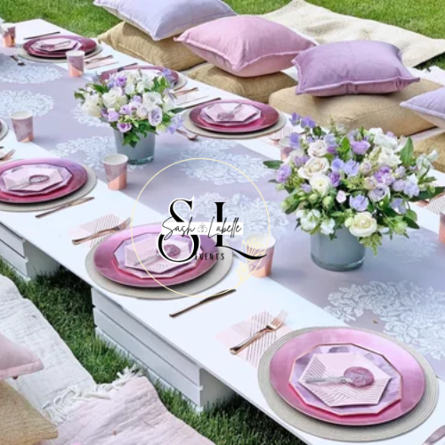 Outdoor picnic table decorated with pink and purple tableware, floral centerpieces, and pastel cushions.
