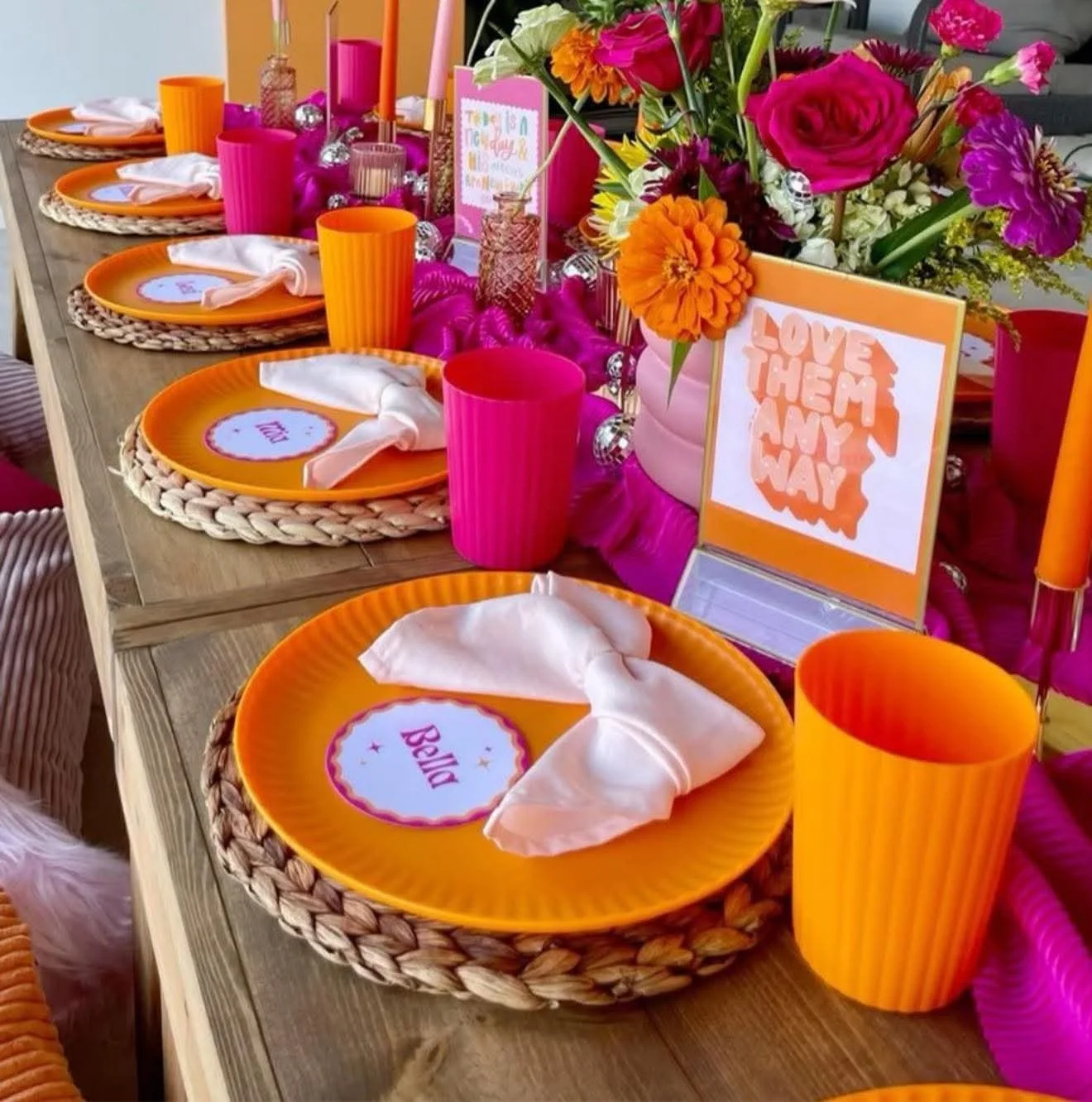 A colorful party table with orange plates labeled with names like 'Bela,' set with white napkins. Bright pink and orange cups, pink candles, and a large flower arrangement with pink, orange, and purple flowers are in the center. Decorations include p