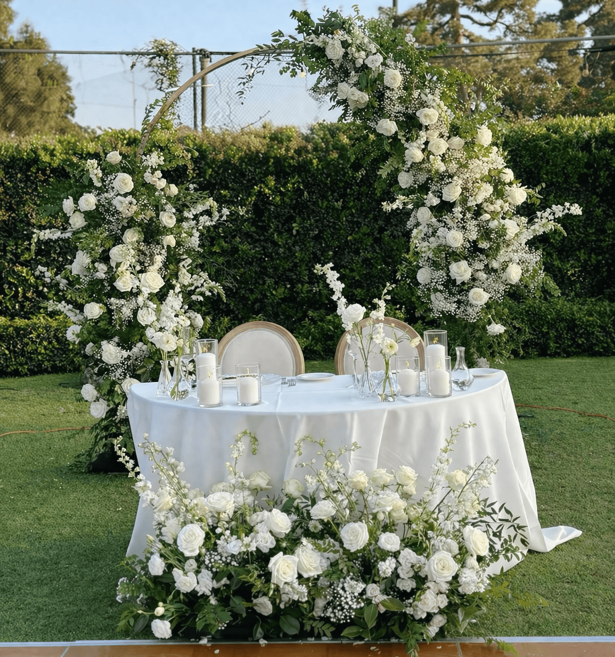 Wedding or event reception table decorated with white flowers and candles, outdoors on green grass with a lush hedge in the background.