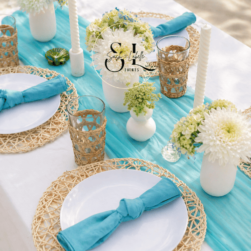 Beachside table setting with white plates, blue napkins, woven placemats, white floral centerpieces, candles, and turquoise table runner.