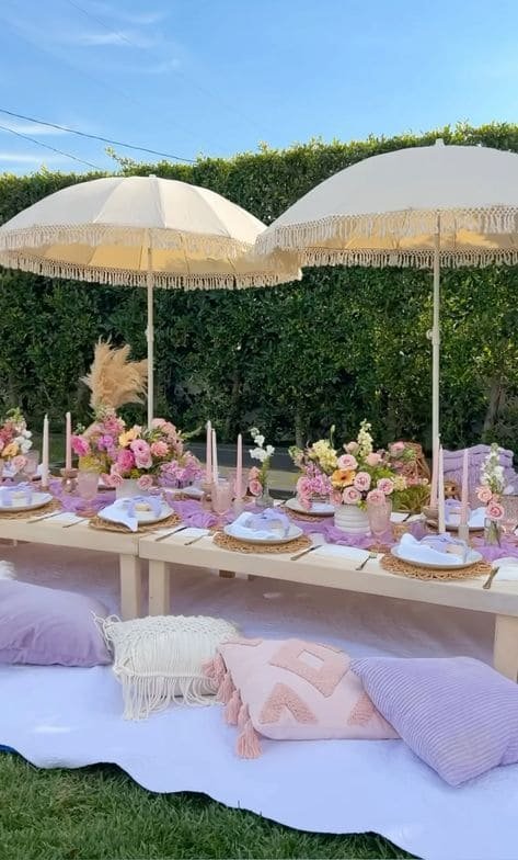 Outdoor table setting with pink and purple flowers, candles, and white plates under two large beige umbrellas, with cushions and blankets on the ground for seating, set against a green hedge and blue sky.