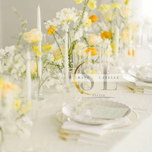 Elegant wedding table setting with white and yellow flowers in glass vases, white plates, gold utensils, and white candles.