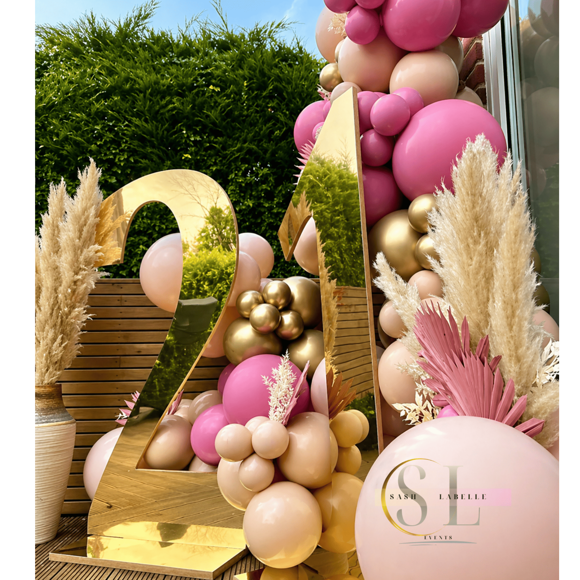 Decorative display featuring a large number 2023 made of gold-colored letters, surrounded by pink, beige, and gold balloons, with pampas grass decorations, set against a wooden fence background, with a promotional sign for an event by Sash Labelle Events.