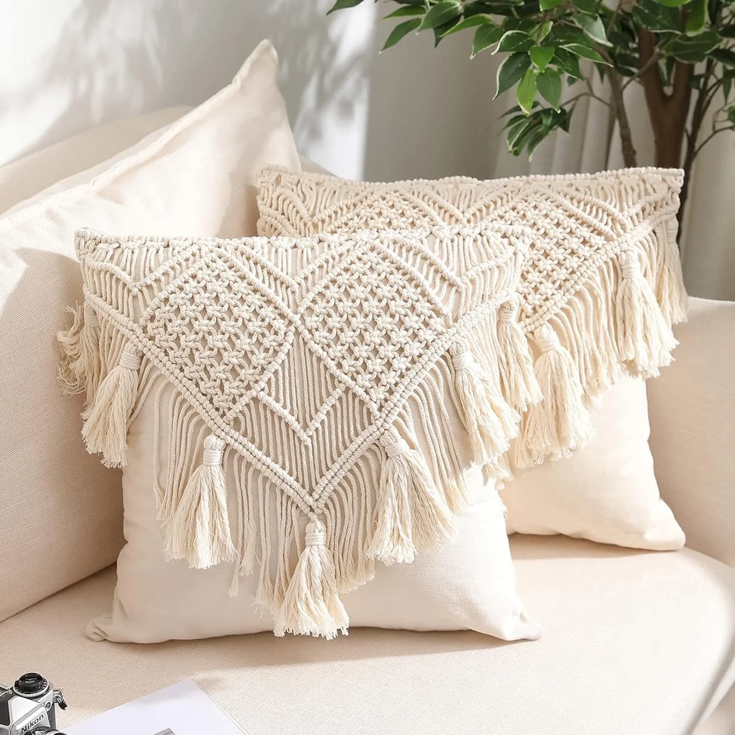 Two cream-colored decorative throw pillows with intricate macrame designs and tassels are on a beige sofa, with a green leafy plant in the background.