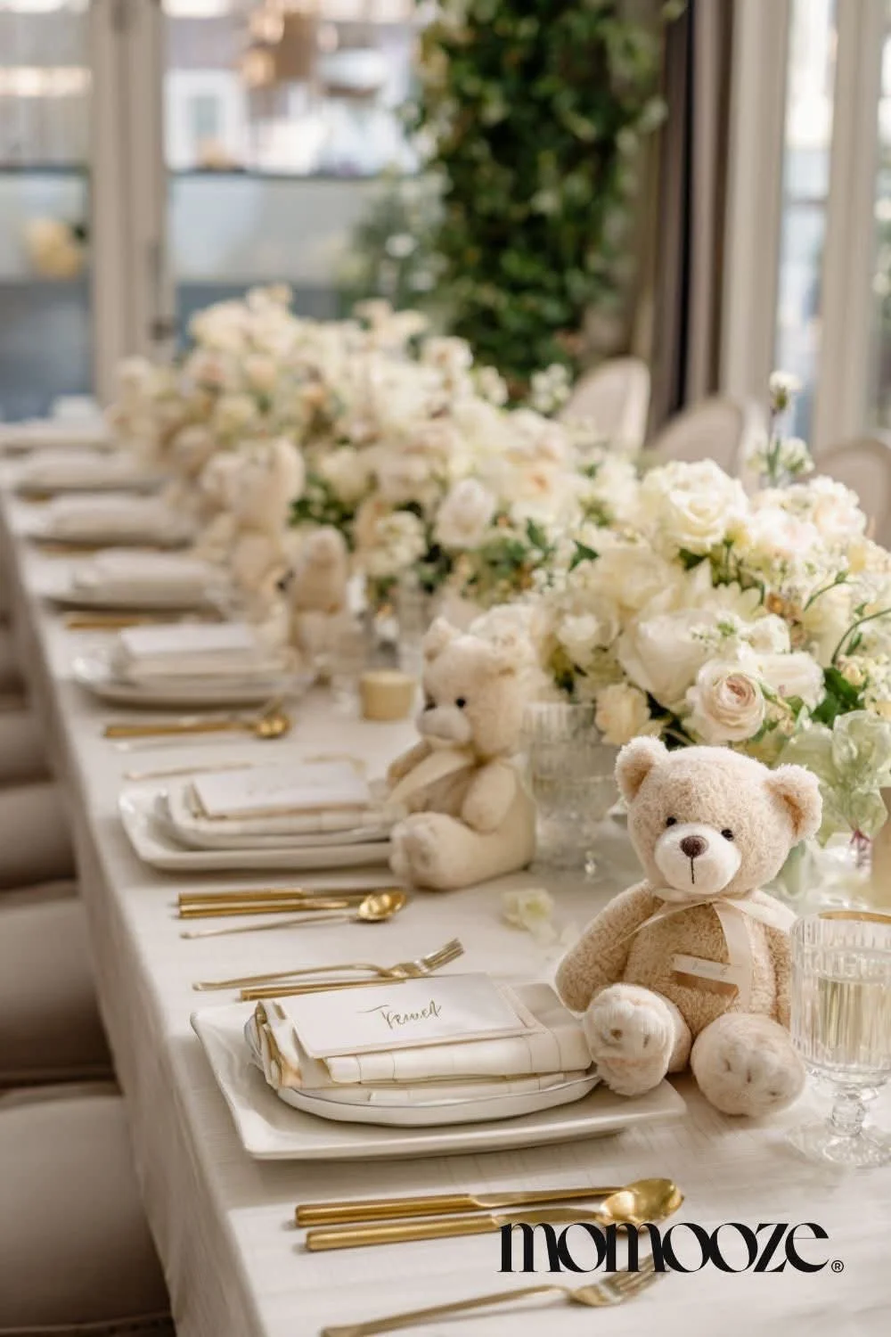A decorated banquet table with white floral arrangements, two teddy bears, gold cutlery, and place cards, set near windows with natural light.