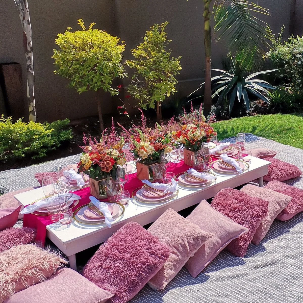 Outdoor dining setup with pink and white cushions, a low white table decorated with pink table runner, floral centerpieces, plates, glasses, and napkins, on a grassy area with trees and plants in the background.