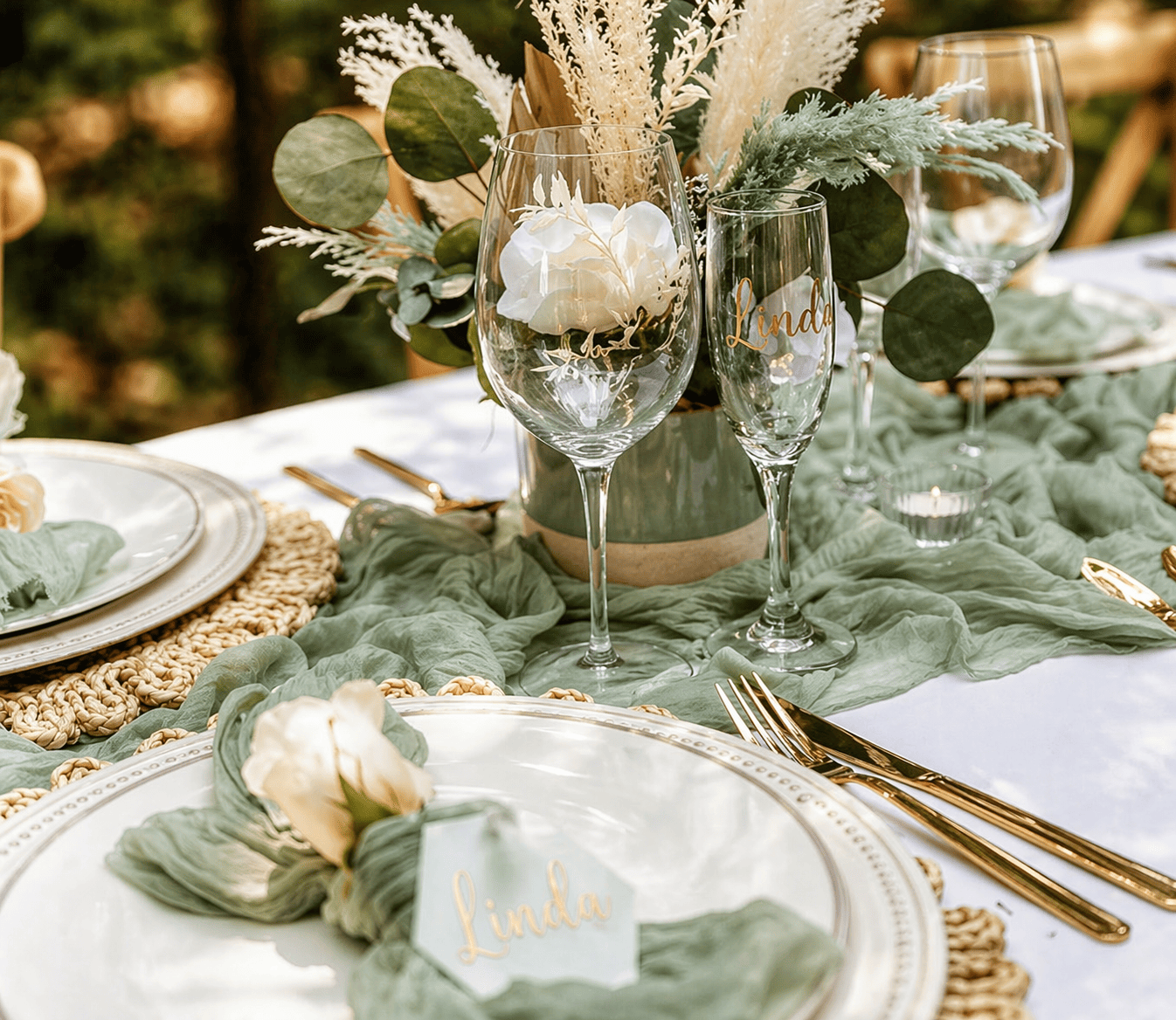 Elegant outdoor table setting with white plates, gold flatware, green cloth napkins, and a floral centerpiece with white flowers and green foliage. Clear wine glasses have gold lettering with the name "Linda."