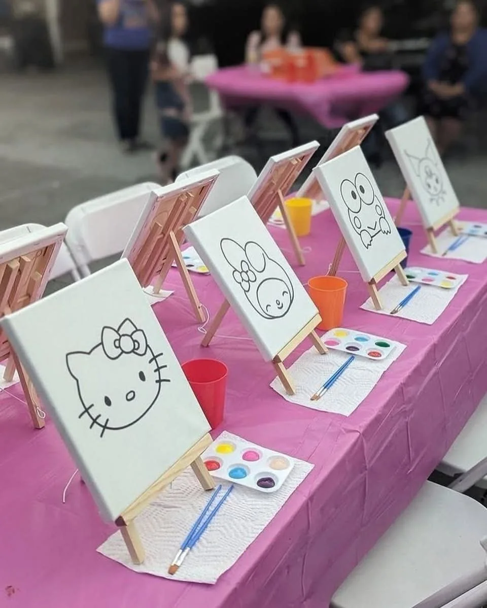 Set up for a children's painting activity with small canvases on easels, featuring Hello Kitty, a bunny, a frog, and a cat drawing, on a pink table with paint palettes, brushes, and colored cups in a group event or party setting.