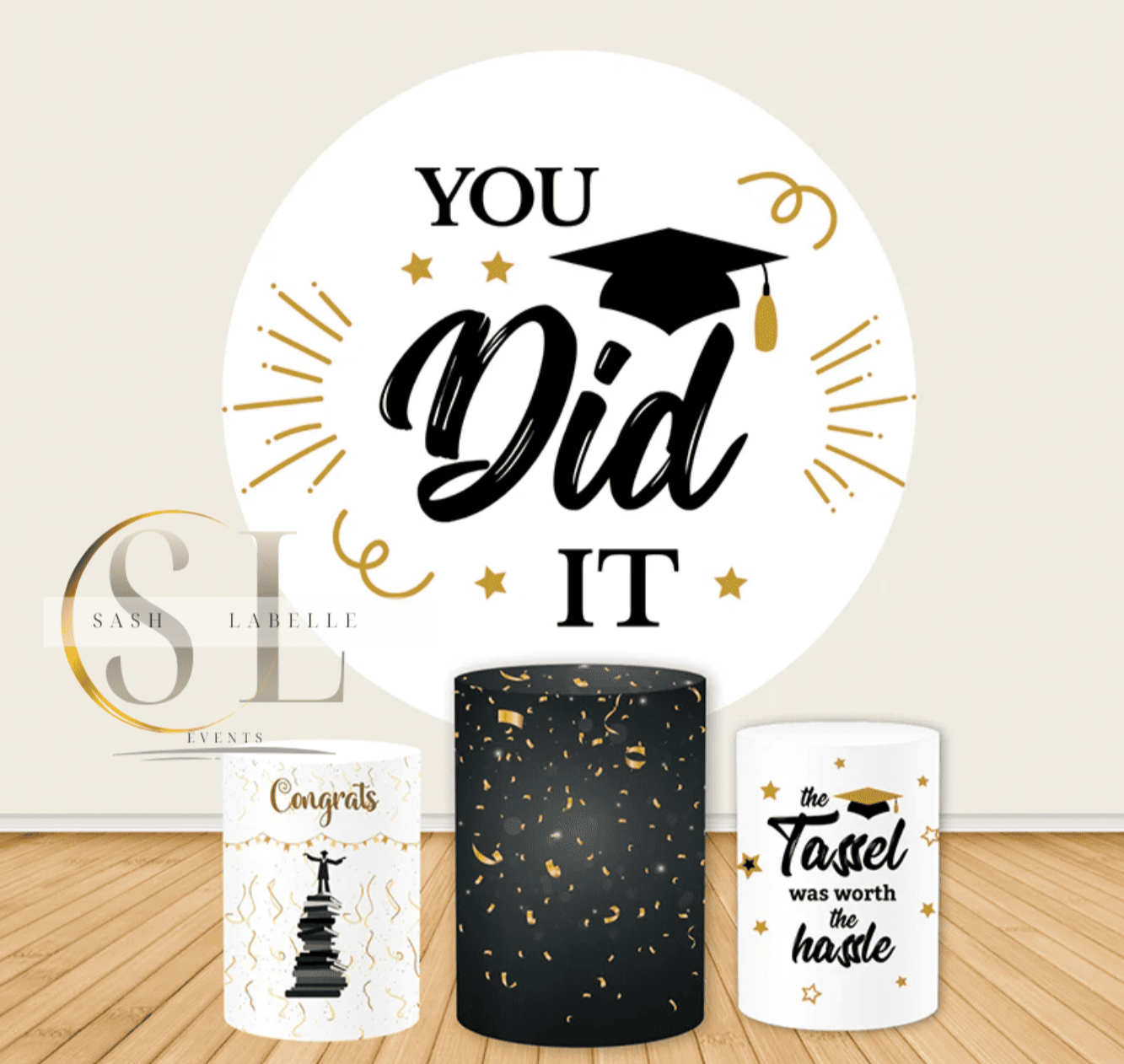 Decorative celebration display with a large circular sign that says "You Did It" with a graduation cap above the word "Did." There are gold stars and confetti accents. In front, there are three cylindrical containers, one white with "Congrats" and a graduation tower graphic, one black with gold confetti, and a white container with a congratulatory message about a towel and the hustle of graduation.