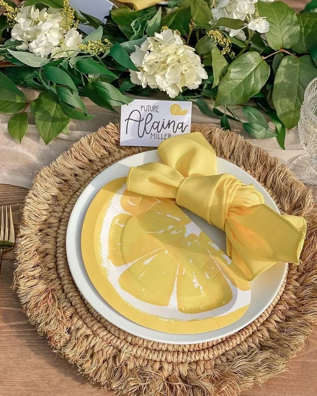 A placemat with a yellow citrus slice pattern, with a matching yellow napkin rolled on a white plate, surrounded by greenery and white flowers, and a card with the name 'Alaina Miller' and 'Future' written on it.